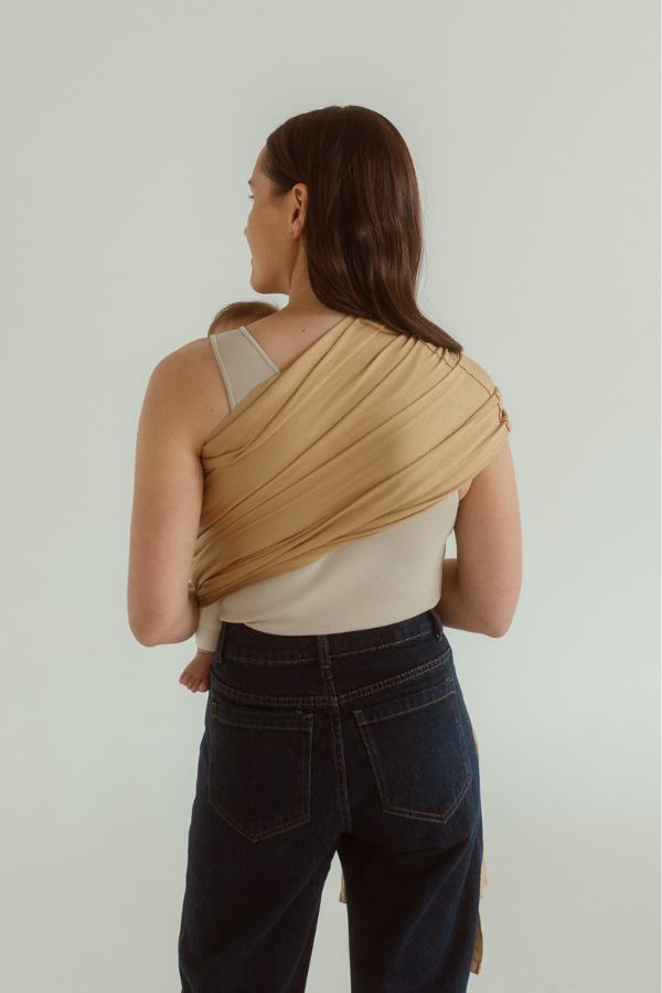 A mother and baby are facing away, showing the back of the Ring Sling Carrier by Chekoh - a soft yellow toned camel colourway fanned evenly across one shoulder and opposite side waist. 