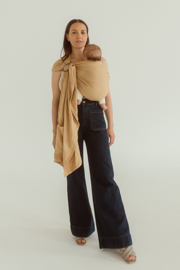 A baby is held close to their mother in a Ring Sling Carrier - it is a soft yellow colour known as Camel, the Rings are visible on the mother's front and a tail of linen and bamboo falls just below her hips revealing a handy pocket too.