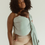 A mother holds her baby close with the help of a linen and bamboo baby ring sling from Chekoh in the colour 'Teal'. A soft green/blue tone.