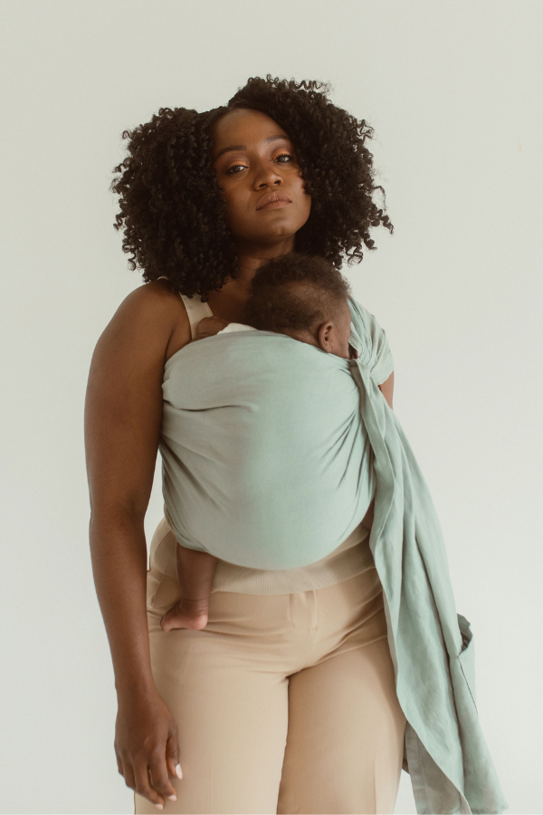 A mother holds her baby close with the help of a linen and bamboo baby ring sling from Chekoh in the colour 'Teal'. A soft green/blue tone.