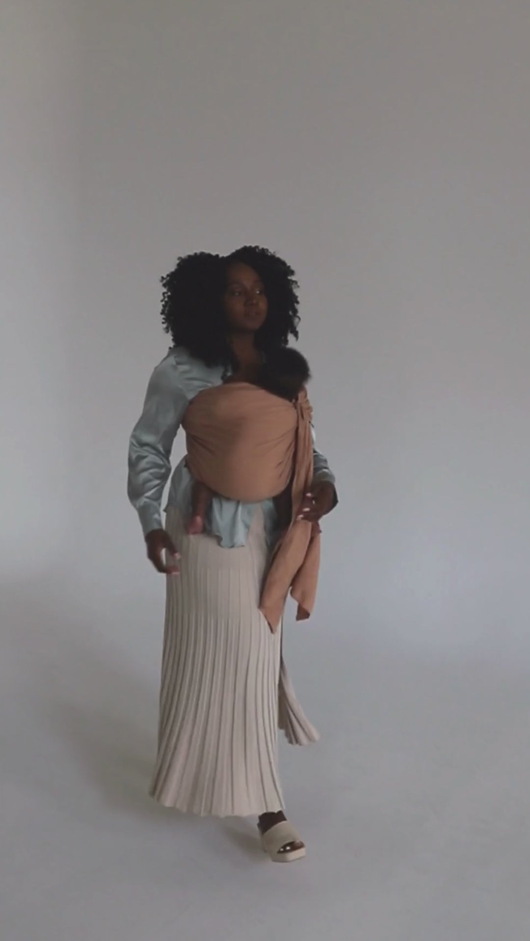 A video of a mother and baby walking across a white backdrop wearing a teal coloured shirt, beige pleated full length skirt and a Terracotta Ring Sling by Chekoh to accent the colour.