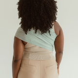 A mother faces away to show off the back of the Teal Ring Sling when worn. The fabric is fanned across her back from one shoulder, down to her opposite side waist. It is a coastal teal tone.