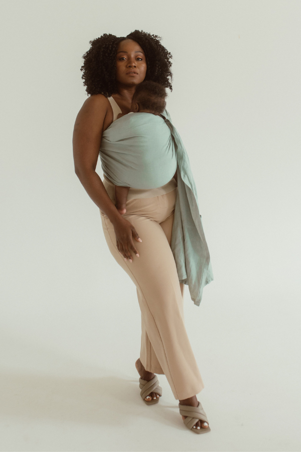 A mother is standing on a white backdrop, wearing the Chekoh Baby Ring Sling Carrier in the shade 'Teal'. A coastal green/blue teal tone - perfect for beachy mamas 