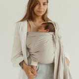 A mother smiles gently, while holding her baby's feet. She is wearing her little one in an Asha grey bamboo and linen Ring Sling Carrier by Chekoh Baby and standing against a white backdrop.