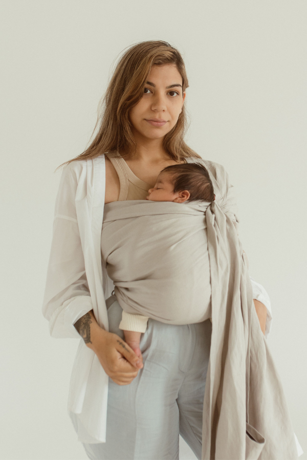 A mother smiles gently, while holding her baby's feet. She is wearing her little one in an Asha grey bamboo and linen Ring Sling Carrier by Chekoh Baby and standing against a white backdrop.