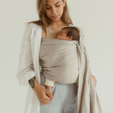 A mother looks down at her little baby, nestled and sleeping in a Chekoh Baby Ring Sling in the colour 'Asha' a soft neutral toned grey.