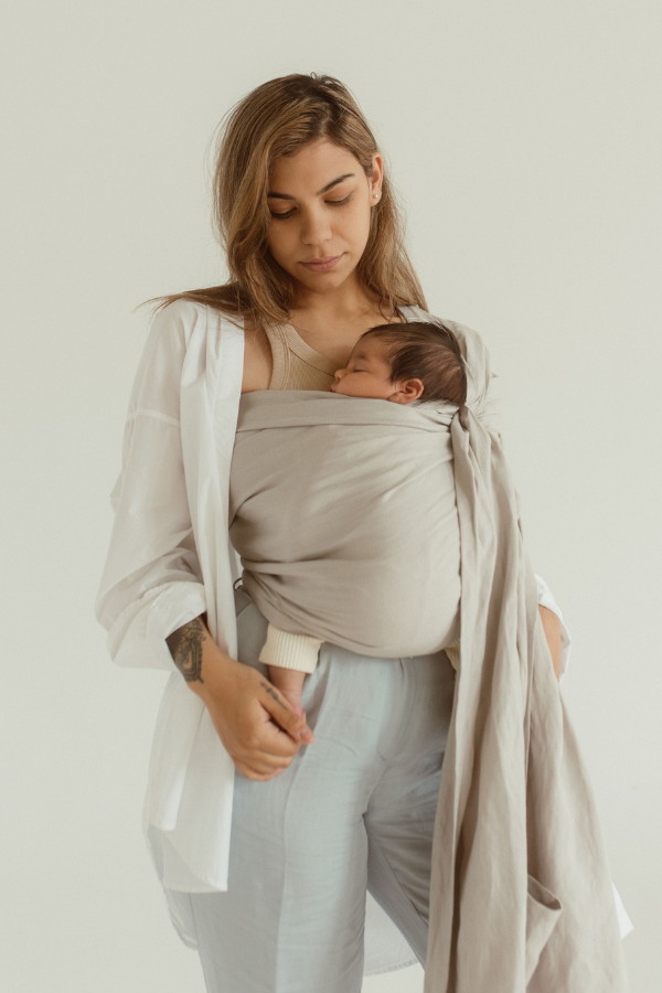 A mother looks down at her little baby, nestled and sleeping in a Chekoh Baby Ring Sling in the colour 'Asha' a soft neutral toned grey.