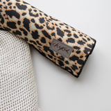 Close up detail of the Chekoh Baby Leopard Print Baby Swaddle Wrap, soft edges have been rolled in to reveal the leopard print. It features colours of brown/orange/tan/cream and black. The the left of this, the corner of the free wash pouch that comes with each swaddle.